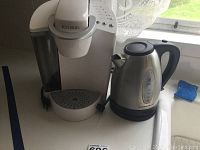 White Keurig coffee maker and stainless steel Hamilton Beach electric kettle on a counter near a window with sheer lace curtain, with power cords visible.