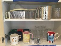 Top kitchen cabinet shelf with a metal box grater, a plastic or glass measuring cup, a stainless steel flour sifter, and clear stackable bowls or cups.