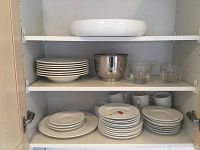 Kitchen cabinet shelf with stacked white ceramic pasta bowl, stacks of plates, white mugs, two clear glass creamer/sugar bowls, and stainless steel bowl.