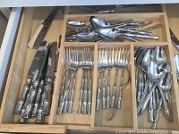 Top view of flatware pieces in wooden drawer organizer showing knives, forks, spoons, and serving spoons arranged in compartments.