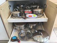 Photo showing upper shelf with multiple stainless steel pots and pans with lids, RoboTwist jar opener and measuring cups stored inside a wooden cabinet.