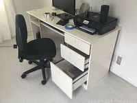 White pressboard desk showing three drawers opened at different levels, black wheeled office chair beside it