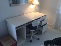 Full view of white MDF desk with three right-side drawers, office chair underneath desk, and white lamp on desk against wall.