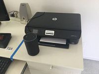Front and side view showing the HP Envy 4500 printer with paper in output tray and Amazon Alexa device on desk near computer keyboard and trackball mouse.