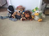 Four stuffed plush toys: two teddy bears, one brown monkey, and one white duck with accessories laid on carpet