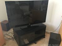 LG 42-inch flatscreen TV mounted on black rolling TV stand with glass doors and caster wheels, situated on beige carpet.