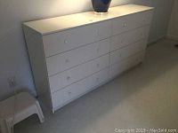 Overall view of white dresser with ceramic lamp on top and small step stool beside it.