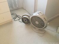 Photo showing two electric fans side by side on floor. Smaller Vornado fan on left, larger Lakewood fan on right.
