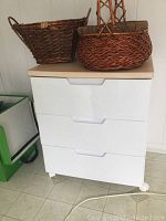 White rolling cabinet with three drawers seen front on with two large woven wicker baskets on top, set on tiled floor.