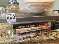 Stack of four cookbooks and perforated baguette pan on kitchen counter with bread proofing baskets on top