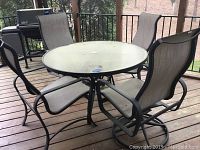 Full view of round glass-top patio table with four surrounding gray mesh fabric patio chairs on wooden porch deck.