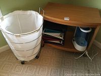 Wooden TV stand with shelf holding three ring binders and white/blue humidifier, next to rolling laundry hamper with white fabric liner and metal frame