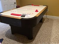 Full view of Turbo air hockey table showing black base, beige trim, and white play surface with red markings and accessories.
