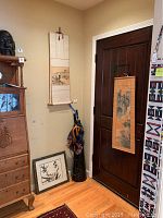 Corner of a room with two silk scrolls hanging on walls, one framed art piece leaning against wall, and an umbrella holder containing four umbrellas