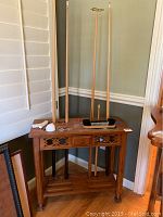 Full view of wooden pool cue table with three pool cues standing behind. Accessories including chalk and brush on top of table.