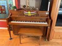 Full front view of upright piano with bench, showing wood finish and music stand with items on top.