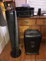 Black Vornado tower fan next to a black paper shredder, both placed on floor next to wooden dresser