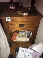 Photo of wooden nightstand showing single drawer with black metal pull, lower shelf with books, surface with slight wear