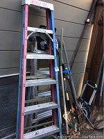 Wide shot showing red and blue Werner fiberglass step ladder along with aluminum multi-position rolling lift ladders stacked behind. Adjacent to the ladders are multiple garden tools including shovels, pitchforks, and rakes, along with a foldable hand truck.