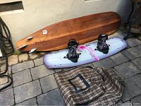 Overhead shot of wooden surfboard, silver-pink Hyperlite wakeboard with bindings, and plaid bag on stone patio