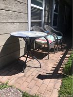 Outdoor mosaic round table with blue-patterned tile top and black metal tripod legs, two plastic woven patio chairs with striped cushions on a brick patio.
