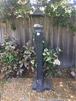 Full front view of the black outdoor propane fire lamp surrounded by garden plants against a wooden fence.