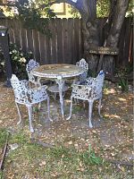 Full view of the table and four iron chairs set in an outdoor setting with some leaves and dirt on the patio surface.