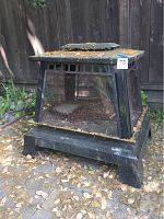 Front view of outdoor fire pit showing mesh sides, flat vented top, and four leg base with surface dirt and pine needles.