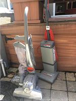 Side and front view of two vintage upright vacuum cleaners: a grey Kirby Heritage II with fabric bag and a chrome front, and a grey Electrolux vacuum with a red power cord.