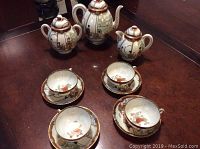 Full set displayed including teapot, creamer, sugar bowl, four cups and saucers, all decorated with detailed geisha scenes on bone china.