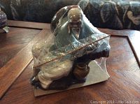 Front angle view of the Mud Man figurine in clear plastic packaging showing details of the figure sitting with staff and pot.