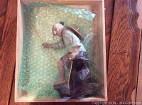Side view of the Chinese Mud Man figurine standing upright inside a wooden box with green bubble wrap padding.