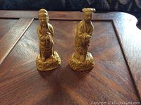 Two small mustard yellow vintage Chinese figures on a wooden table. Both figures wear traditional robes and stand on decorated bases.