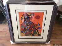 Framed Norval Morrisseau print wrapped in protective plastic, wooden frame visible, artwork depicts colorful abstract figures on orange background.