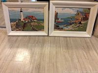 Two vintage paint by numbers paintings displayed side by side on the floor showing lighthouse and dock scenes in white frames.