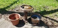 Wide outdoor image showing painted cement bird bath flanked by two plastic pots and metal chimes on ground