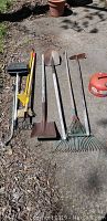 Full view of garden tools lined up on pavement including shovels, rakes, dustpan, and gas can