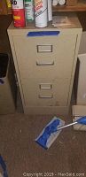 Beige metal 2-drawer file cabinet appears closed with cleaning mop in front, located in storage area.