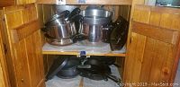Cabinet shelf with stacked stainless steel pots and pans, some with copper bottoms, and non-stick frying pans below
