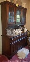 Full front view of the Bassett wood 2-piece hutch showing upper glass cabinets, lower storage cabinets, drawers, and some decorative items on top and surface.