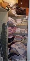 Shelves with folded bed linens and towels in white, pink, and beige tones with ironing board visible