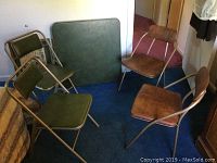 Full view of the card table with green vinyl top and four folding chairs, two with green padded vinyl seats and backs, two with wood-tone seats and backs, all metal framed.