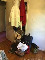 Room corner showing two ladies coats hung on door (beige London Fog and red Forecaster) with assorted handbags and purses below, and several pieces of luggage on the floor.