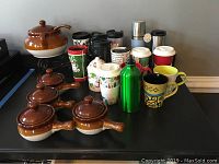 Five ceramic brown and cream soup crocks with lids and spoons, alongside assorted coffee mugs and travel cups in various colors and designs, including green, red, black, white, and metallic finishes.