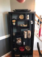 Black shelving unit showing three black metal mesh bread trays on top and middle shelves. Various ceramic, glass and other kitchen decor arranged within compartments.