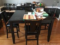 Wide view of black square wooden dining table with four matching chairs around it. Table top has some items placed on it, and surface scratches visible.