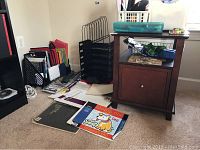 Photo showing black wire and plastic organizers, colored folders and office supplies on floor near wooden side cabinet