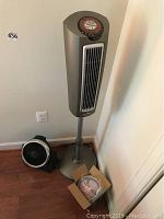 Silver tall tower fan with control panel on top, black compact circular fan on floor, box containing white small circular fan with blades.