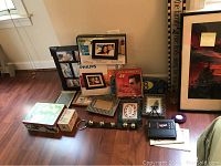 Overview of lot showing various framed art, Coby and Philips digital photo frames, sleigh bells, decorative tins, and other small items arranged on the floor.