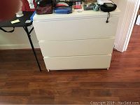 White laminated wooden dresser photographed from the front showing all three closed drawers and surface with various items on top.